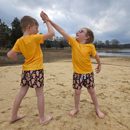 Two young boys wearing popcorn swim trunks and yellow rash guards high fiving each other at the beach. 